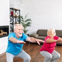 An older man and woman demonstrate how to do a Kua Squat. They hold their arms out front and parallel to the floor. They bend from their knees and keep their backs as straight as possible.